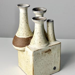 May include: A white ceramic vase with four openings, each with a different shape and size. The vase has a speckled texture and a brown band around the bottom of the largest opening.