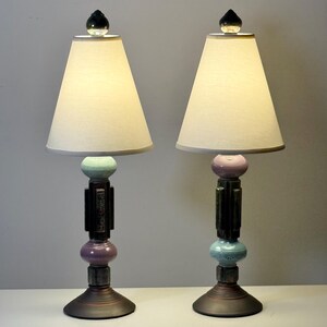 May include: Pair of decorative table lamps with off-white tapered shades. The bases feature a dark, geometric design with pastel blue and purple accents. Each lamp is topped with a clear, faceted finial.
