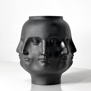 May include: A matte black ceramic vase featuring four sculpted faces. The faces are arranged around the vase, each with detailed features. The vase has a rounded body and a slightly flared opening, suitable for home decor.