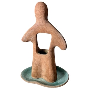 May include: A terracotta ceramic sculpture of a person with a cutout in the middle, standing on a green ceramic base.
