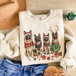May include: A cream-colored sweatshirt with a Christmas-themed design featuring four German Shepherd dogs wearing Santa hats and reindeer antlers. The dogs are surrounded by red and green Christmas decorations.