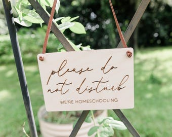 May include: A rectangular light-colored wooden sign hanging from a brown leather cord. The sign features the engraved text "please do not disturb" in a cursive script font, followed by the text "WE'RE HOMESCHOOLING" in a simple sans-serif font.