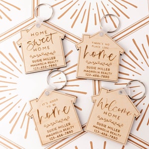 May include: Four wooden house-shaped keychains with different sayings about home. Each keychain has a silver key ring and features the text "Susie Miller Magnolia Realty 123-456-7890". The sayings are "Home Sweet Home", "There's No Place Like Home", "It's Good To Be Home", and "Welcome Home".