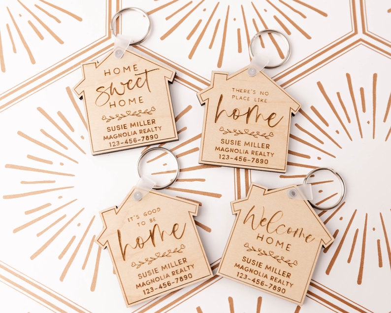 May include: Four wooden house-shaped keyrings with different sayings about home. Each keyring has a silver key ring and features the text "Susie Miller Magnolia Realty 123-456-7890". The sayings are "Home Sweet Home", "There's No Place Like Home", "It's Good To Be Home", and "Welcome Home".