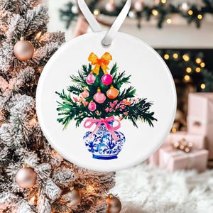 May include: A white ceramic ornament featuring a hand-painted watercolor illustration of a Christmas tree in a blue and white porcelain vase. The tree is decorated with pink and orange ornaments and a yellow bow.