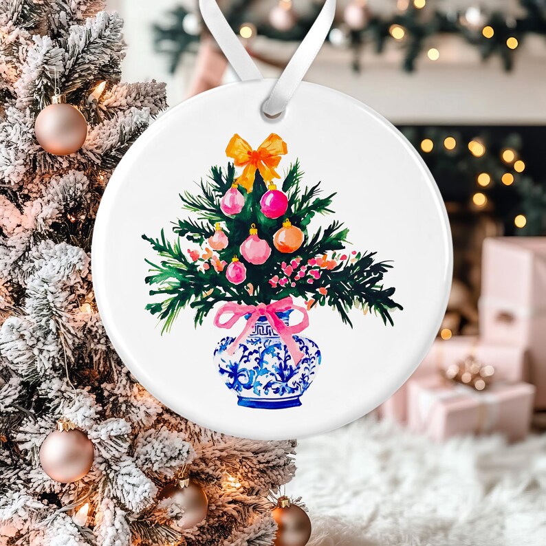 May include: A white ceramic ornament featuring a hand-painted watercolor illustration of a Christmas tree in a blue and white porcelain vase. The tree is decorated with pink and orange ornaments and a yellow bow.