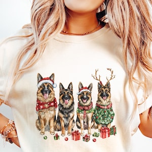 May include: A white t-shirt with a Christmas-themed design featuring four German Shepherd dogs wearing Santa hats and reindeer antlers. The dogs are surrounded by presents and green and red ornaments.