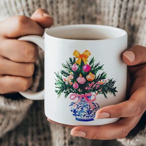 May include: White ceramic mug with a hand-painted design of a Christmas tree in a blue and white vase. The tree is decorated with pink and yellow ornaments and a yellow bow.