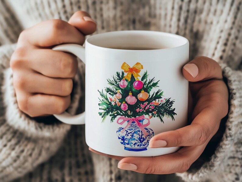 May include: White ceramic mug with a hand-painted design of a Christmas tree in a blue and white vase. The tree is decorated with pink and yellow ornaments and a yellow bow.