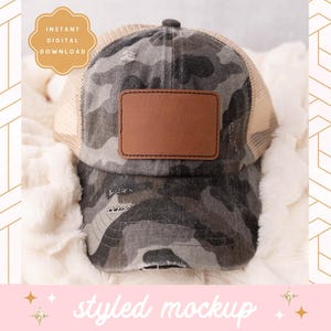 Könnte beinhalten: Eine Camouflage-Trucker-Kappe mit einem braunen Leder-Patch auf der Vorderseite. Die Kappe liegt auf einer weißen, flauschigen Oberfläche.