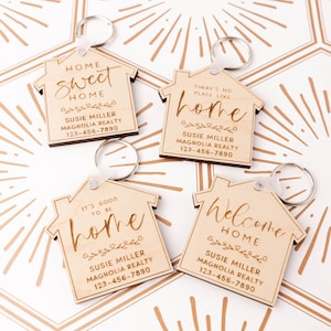 May include: Four wooden house-shaped keyrings with different sayings about home. Each keyring has a silver key ring and features a different saying: "Home Sweet Home", "There's No Place Like Home", "It's Good To Be Home", and "Welcome Home". Each keyring also includes the name "Susie Miller" and "Magnolia Realty" with a phone number.