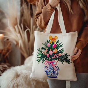 May include: A white canvas tote bag with a colorful illustration of a Christmas tree in a blue and white porcelain vase. The tree is decorated with pink, yellow, and green ornaments.