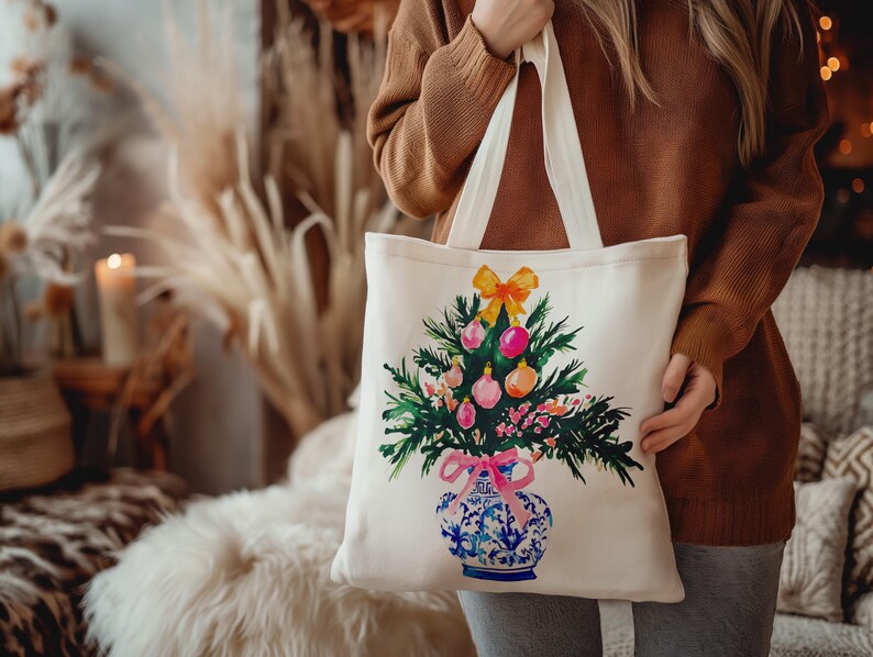 May include: A white canvas tote bag with a colorful illustration of a Christmas tree in a blue and white porcelain vase. The tree is decorated with pink, yellow, and green ornaments.