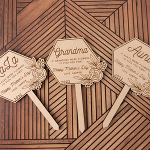 May include: Three wooden hexagonal garden stakes with floral designs and personalized messages for Mother's Day. The stakes read "LaLa", "Grandma", and "Aunt" with a personalized message and the year 2022.