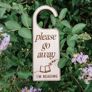 May include: A light-colored wooden door hanger with a hook top. The text "please go away" is engraved in a cursive font above a line drawing of an open book with a butterfly. The text "I'M READING" is engraved in a bold, sans-serif font at the bottom.