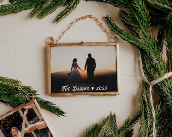 May include: A rectangular gold-toned metal frame with a hanging chain containing a photograph of a couple holding hands at sunset. The text "The Barons ♥ 2023" is printed in white at the bottom of the glass. A second similar frame is partially visible.