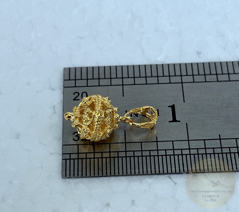 May include: Gold-toned pendant charm with a detailed, intricate design. The charm is approximately 1 inch in diameter and features a loop for attaching to a necklace or bracelet.