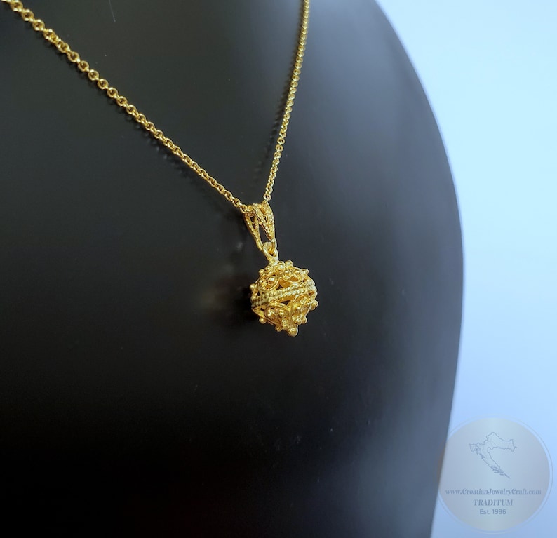 May include: A gold chain necklace with a delicate, intricate gold pendant in the shape of a sphere. The pendant has a textured surface and appears to be made of multiple interwoven strands.