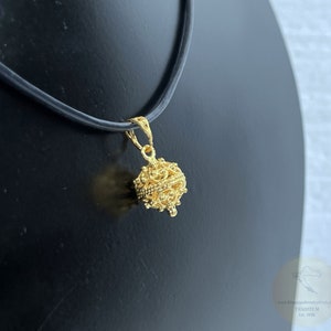 May include: A gold-colored pendant necklace with a detailed, ornate, spherical charm. The charm is suspended from a black leather cord.