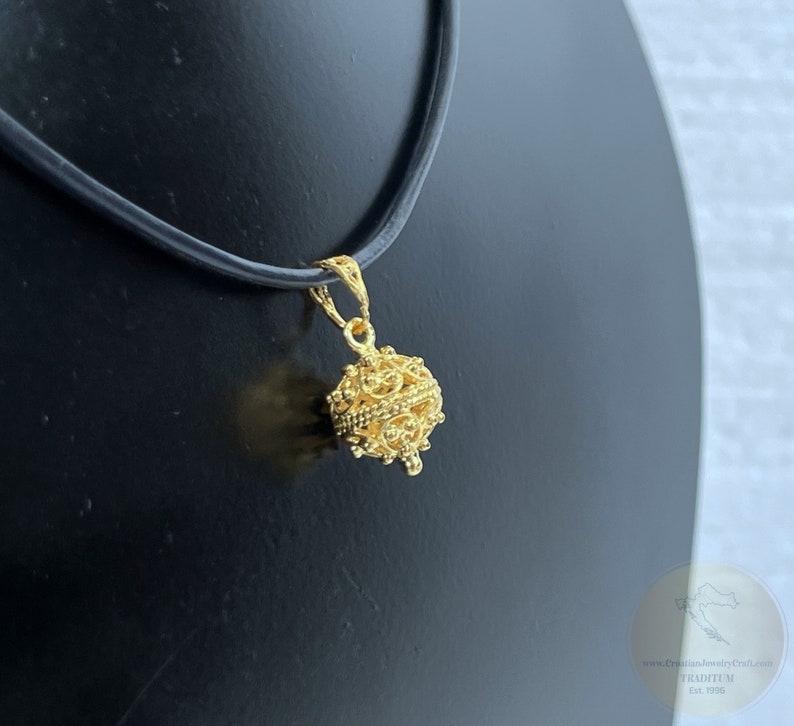May include: A gold-colored pendant necklace with a detailed, ornate, spherical charm. The charm is suspended from a black leather cord.