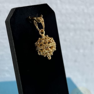 May include: A gold pendant in the shape of a round, intricate cage with a small loop at the top for attaching to a chain. The pendant is on a black display stand.