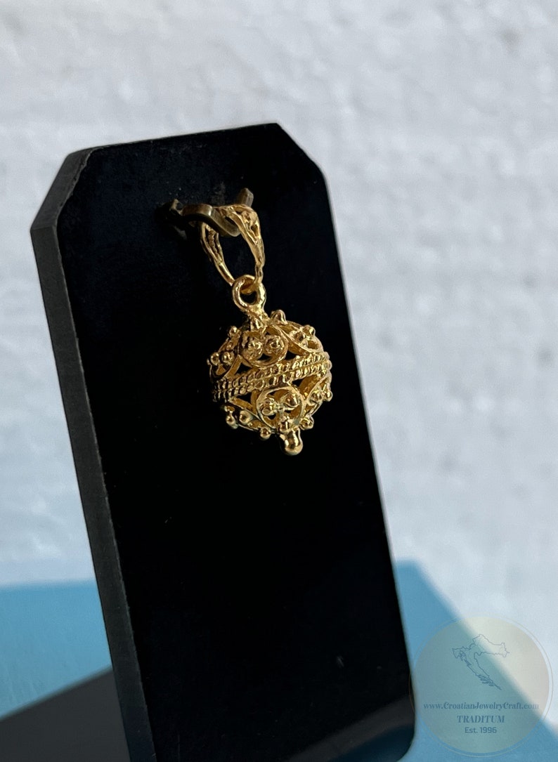 May include: A gold pendant in the shape of a round, intricate cage with a small loop at the top for attaching to a chain. The pendant is on a black display stand.