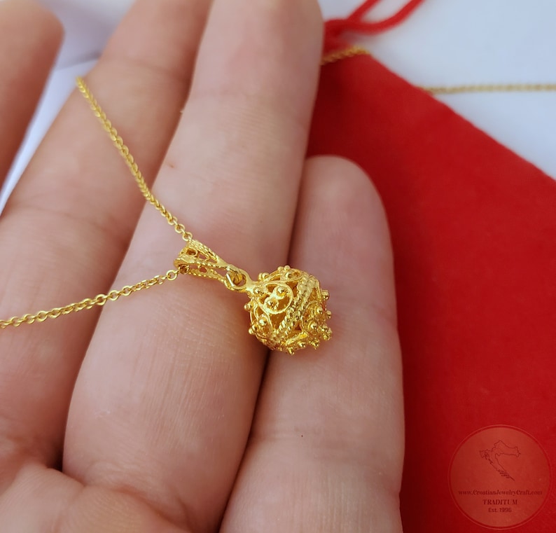 May include: A gold chain necklace with a delicate, ornate pendant. The pendant is a small, round ball with intricate designs and a textured surface.