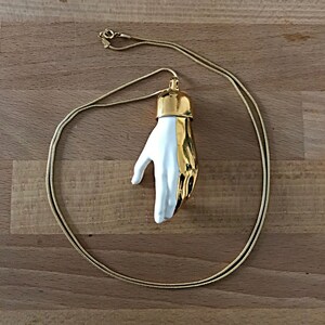 May include: A gold chain necklace with a white ceramic hand pendant. The hand is stylised with a gold cuff around the wrist.