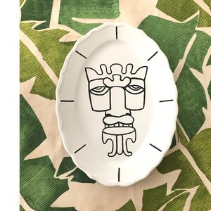 May include: A white ceramic plate with a black and white line drawing of a tiki mask on it. The plate is on a green and white patterned surface.