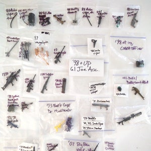 May include: A collection of plastic action figures and accessories in clear plastic bags. The bags are labeled with the year and name of the item. The items are from the G.I. Joe toy line. '98+ UP GI Joe Acc. is written on a white card.