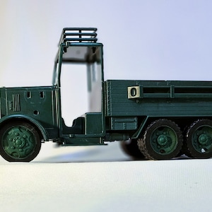May include: A dark green toy truck with six wheels and a flatbed. The truck has a detailed cab with a roof rack and a small number plate. The wheels and other details are also dark green.