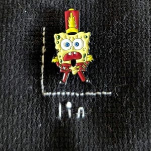 May include: A yellow, red, and gold enamel pin of Spongebob Squarepants in a marching band uniform. The pin is on a dark gray textured fabric. The pin has blue eyes and a red mouth. The text "lin" is stitched in white thread below the pin.