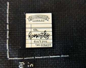 May include: A rectangular white enamel pin shaped like a library card with black text reading HOGWARTS LIBRARY, Oh, honestly, don't you two READ? The pin is positioned next to a white ruler marking 2 inches, showing the pin is under 2 inches tall.