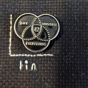 Peut inclure: Une épingle en argent avec un motif en émail noir. Le motif est un diagramme de Venn avec trois cercles qui se chevauchent. Les cercles sont étiquetés "Vie", "Univers" et "Tout". Le centre du diagramme contient le nombre "42".