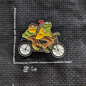 May include: A green and yellow frog couple riding a tandem bicycle with a red heart above them. The bicycle has white wheels and black handlebars.