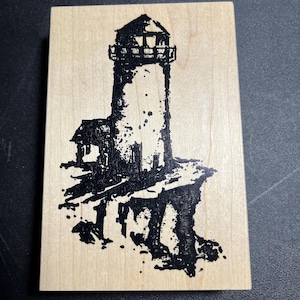 May include: A wooden stamp featuring a black ink illustration of a lighthouse. The lighthouse is depicted with a detailed tower and a small structure at its base, set against a light wood background.