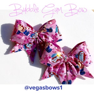 May include: Two pink cheer bows with a Bubble Yum theme. The bows are decorated with rhinestones and feature a pink and blue cartoon character with a bubble gum.