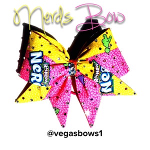 May include: A pink and yellow cheer bow with a Nerds candy wrapper design. The bow is decorated with rhinestones and has a red elastic band.
