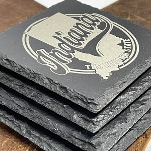 May include: Set of four black slate coasters with a silver engraved design. The design features the state of Indiana with the text "Indiana" and "The Hoosier State".