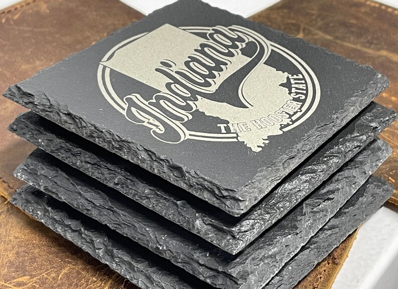 May include: Set of four black slate coasters with a silver engraved design. The design features the state of Indiana with the text "Indiana" and "The Hoosier State".