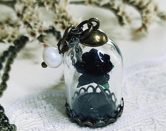 May include: A pendant featuring a miniature black rose encased in a clear glass dome with a bronze-toned metal base and cap. The piece hangs from a bronze-toned chain and is accented with a single white pearl bead attached to the top jump ring.