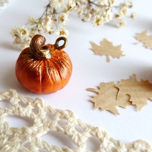 Puede incluir: Cuatro pequeñas calabazas de color naranja y dorado con un tallo dorado y un lazo para colgar. Las calabazas están sobre una superficie blanca con hojas de papel marrón y un encaje blanco de ganchillo.