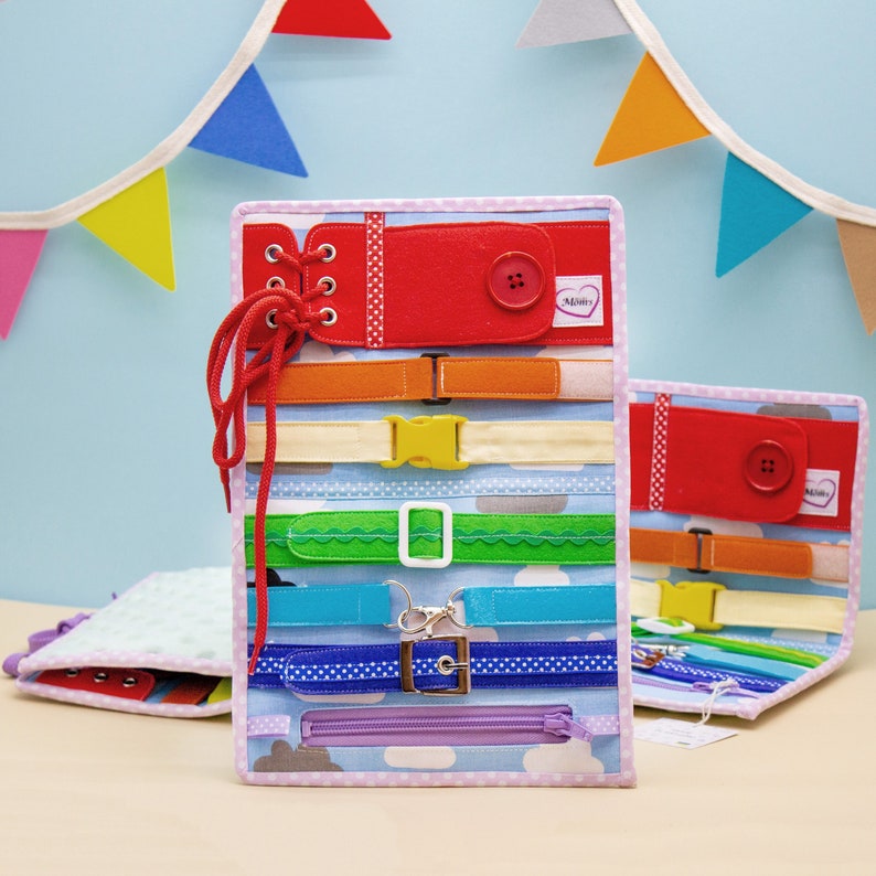Puede incluir: Un tablero de actividades de tela rosa y blanco con una variedad de texturas y cierres para ni&ntilde;os. El tablero presenta una cremallera, cordones, botones, hebillas y una variedad de cintas de colores. El tablero est&aacute; etiquetado como "Mommy".