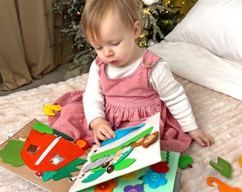 May include: A child in a pink dress plays with a felt activity book. The book has different pages with various textures and colors, including a red tent, a green tree, and a blue flower.