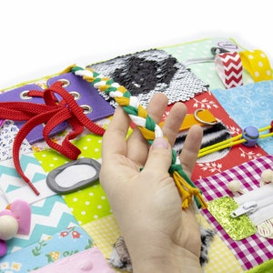 May include: A multi-colored fabric activity mat with various textures and features for babies and toddlers. It includes a zipper, laces, buttons, a buckle, a squeaky toy, and a variety of fabrics.