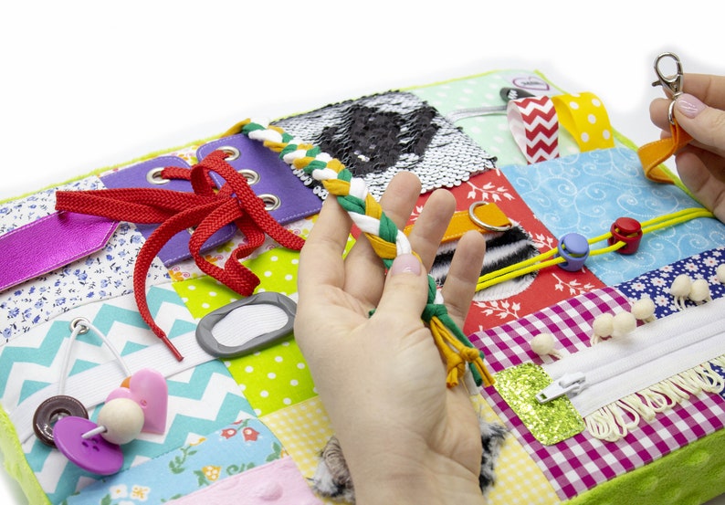 May include: A multi-colored fabric activity mat with various textures and features for babies and toddlers. It includes a zipper, laces, buttons, a buckle, a squeaky toy, and a variety of fabrics.