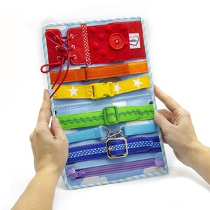 Puede incluir: Un tablero de actividades de colores arco&iacute;ris con varios cierres, incluyendo una cremallera, una hebilla, un cierre de velcro y un cierre de encaje. El tablero est&aacute; hecho de fieltro y tiene un borde a rayas azul y blanco.