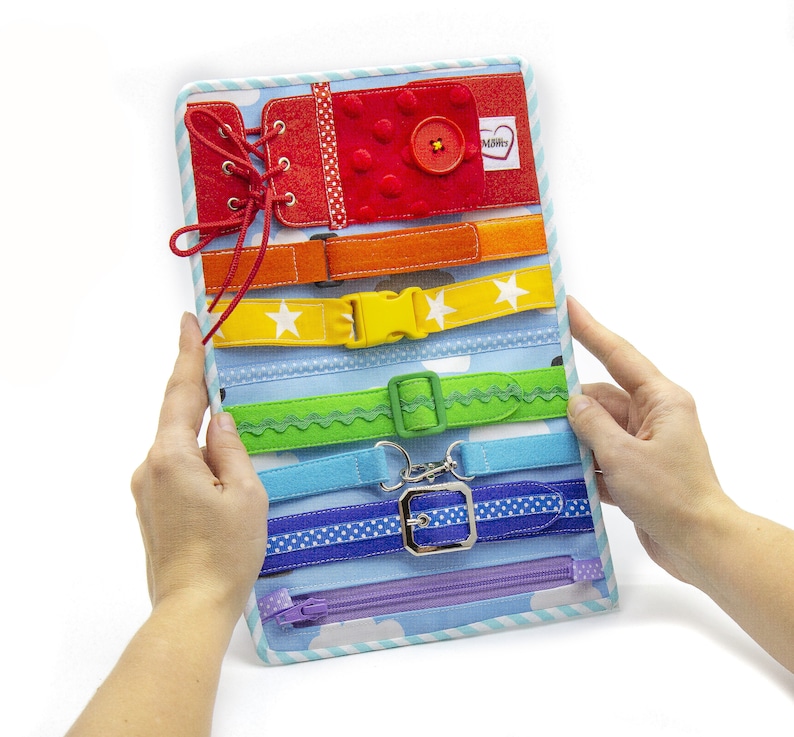 Puede incluir: Un tablero de actividades de colores arco&iacute;ris con varios cierres, incluyendo una cremallera, una hebilla, un cierre de velcro y un cierre de encaje. El tablero est&aacute; hecho de fieltro y tiene un borde a rayas azul y blanco.