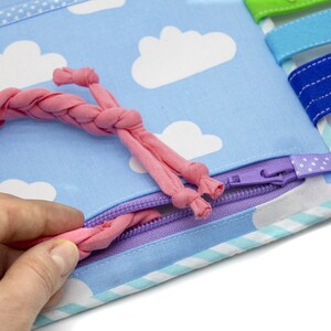 Puede incluir: Un lazo de tela rosa se est&aacute; tirando a trav&eacute;s de una cremallera morada en un libro de tela azul con nubes blancas impresas en &eacute;l. El libro tiene un borde a rayas azul claro y blanco.