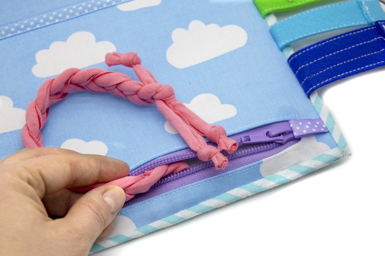 Puede incluir: Un lazo de tela rosa se est&aacute; tirando a trav&eacute;s de una cremallera morada en un libro de tela azul con nubes blancas impresas en &eacute;l. El libro tiene un borde a rayas azul claro y blanco.